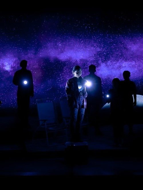 A group of actors stand with flashlights on a dark stage, backlit by an image of the Milky Way